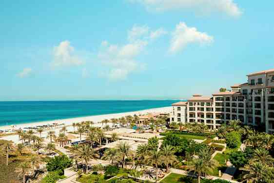 Utsikt over et feriested ved sjøen med en bygning, grøntområde og palmer i forgrunnen. (AI-generert tekst)