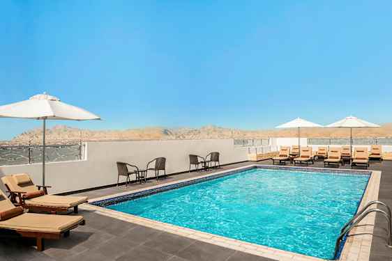 Et utendørs basseng omgitt av solsenger med parasoller og sittegrupper, med utsikt over fjell og en klar blå himmel. (AI-generert tekst)