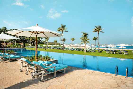 Et basseng med solsenger og parasoller i et utendørsmiljø, omgitt av palmer og plen. (AI-generert tekst)