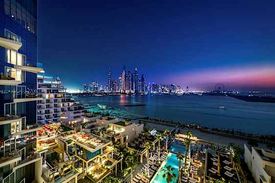 Utsikt over et moderne bymiljø ved skumring med skyskrapere, hav og en bygning med opplyste terrasser og bassengområde i forgrunnen. (AI-generert tekst)