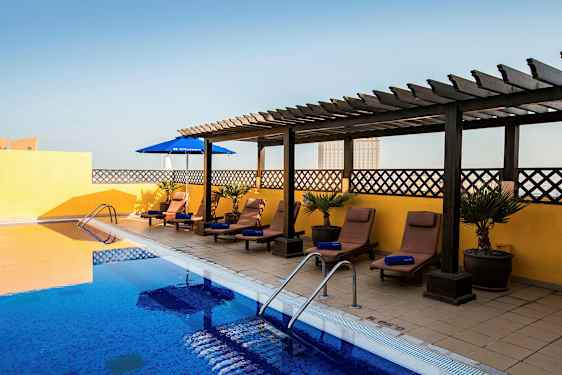 Et utendørsbasseng med blått vann omgitt av solsenger og parasoller. Solsengene står på et belagt gulv med en gul vegg og trepergola i bakgrunnen. (AI-generert tekst)