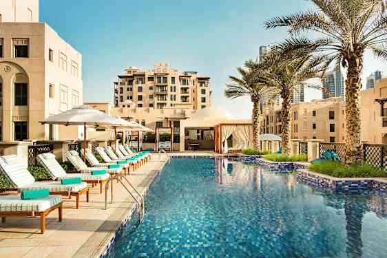 Et utendørs svømmebasseng omgitt av solsenger med parasoller og palmer ved siden av bygninger. (AI-generert tekst)