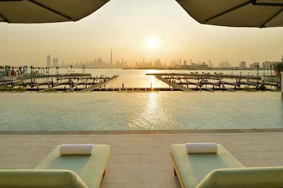 Utsikt fra et evighetsbasseng mot en marina og en bysilhuett under en solnedgang, med to solsenger i forgrunnen. (AI-generert tekst)