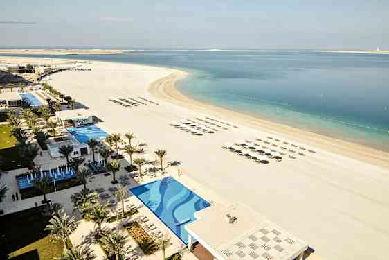 Bildet viser en strand med parasoller og solsenger i rette rader, ved siden av et basseng og et hotellområde omgitt av palmer. (AI-generert tekst)