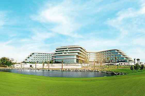 Et moderne hotell med flervåningsbygg omgitt av grønne områder og en dam. (AI-generert tekst)