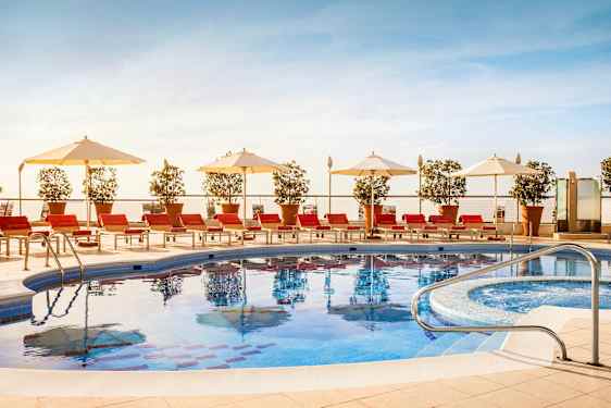 Et utendørs svømmebasseng omgitt av røde solsenger, hvite parasoller og pottede planter. (AI-generert tekst)