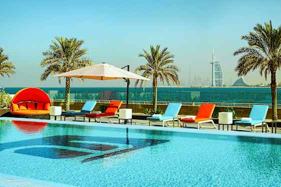 Et basseng med solsenger og parasoller, omgitt av palmer med havutsikt og en bysilhuett i bakgrunnen. (AI-generert tekst)