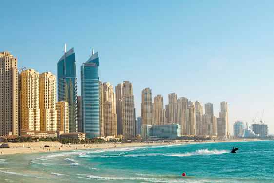 Bilde av høyhus ved en strand med klart blått vann, en person er på en jetski nær stranden. (AI-generert tekst)