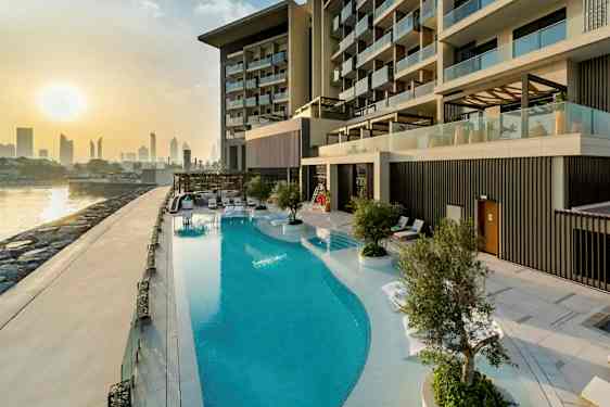 Bilde av et luksuriøst hotellbasseng ved en bygning med moderne balkonger. I bakgrunnen sees en bysilhuett og solnedgang. (AI-generert tekst)