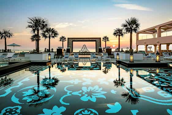 Bilde av et basseng med dekorativt mønster og omkringliggende solsenger, palmer og solnedgang i bakgrunnen. (AI-generert tekst)