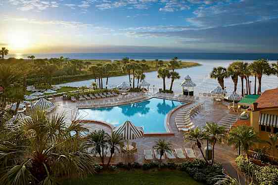 Bilde av et bassengområde med solsenger og parasoller, omgitt av palmer og en strand i bakgrunnen. (AI-generert tekst)