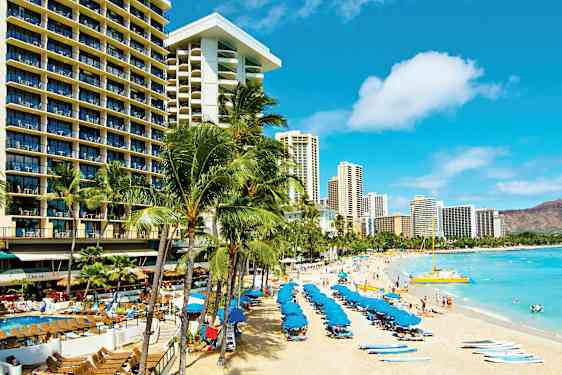 En sandstrand med solsenger og blå parasoller, omgitt av høye hotellbygninger og palmer. (AI-generert tekst)
