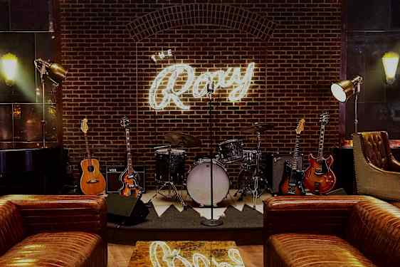 En scene med musikkinstrument som trommer og gitarer foran en murvegg med et lysende skilt som sier "The Roxy". (AI-generert tekst)