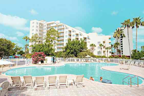 Et utendørs svømmebasseng foran en hvit bygning omgitt av trær og solsenger, med mennesker i vannet og en klar blå himmel over. (AI-generert tekst)