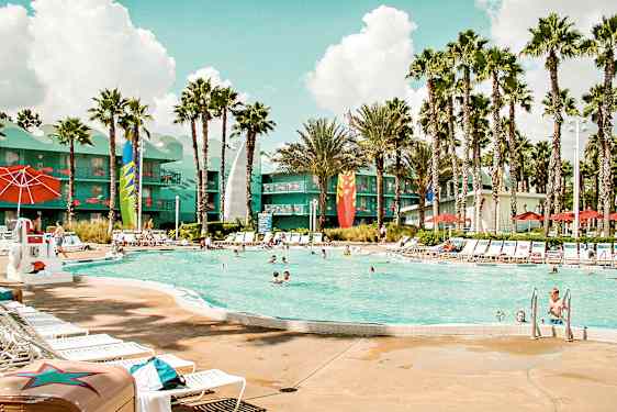 Et utendørs basseng omgitt av solsenger, parasoller og palmer. I bakgrunnen sees en bygning med fargerike detaljer. (AI-generert tekst)