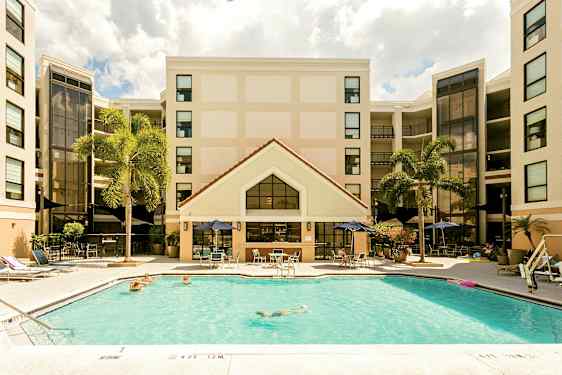 Bilde av et hotell med et basseng i forgrunnen. Personer svømmer og slapper av ved bassengområdet. Hotellfasaden er synlig i bakgrunnen med palmer i nærheten. (AI-generert tekst)