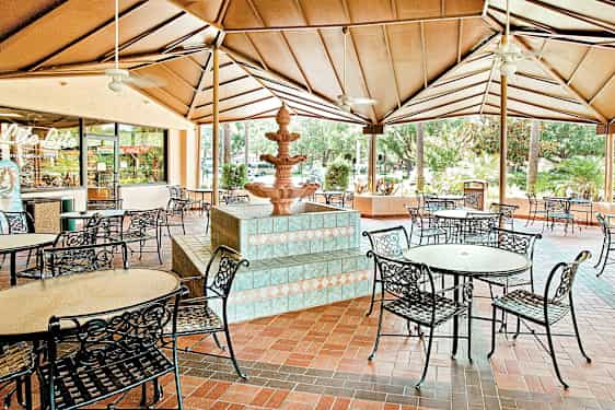 En overbygd uteplass med runde bord og metallstoler og en fontene i midten. (AI-generert tekst)