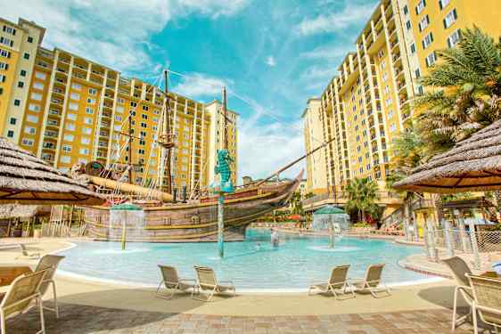 Et basseng omgitt av solsenger og gule flerboligbygg med et piratskip i midten. (AI-generert tekst)