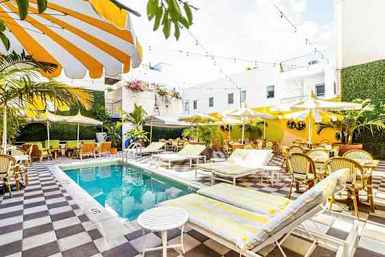 Et lyst og fargerikt utendørs svømmebasseng omgitt av solsenger, parasoller og sitteplasser med grøntområder og hvite bygninger. (AI-generert tekst)