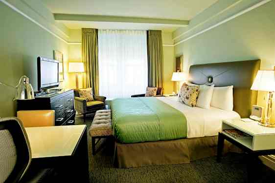 Et hotellrom med en stor seng, et skrivebord og en TV. Rommet er innredet i grønne toner og har store vinduer. (AI-generert tekst)