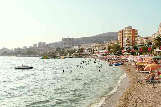 En strand med svømmende mennesker og parasoller, samt bygninger i bakgrunnen. (AI-generert tekst)