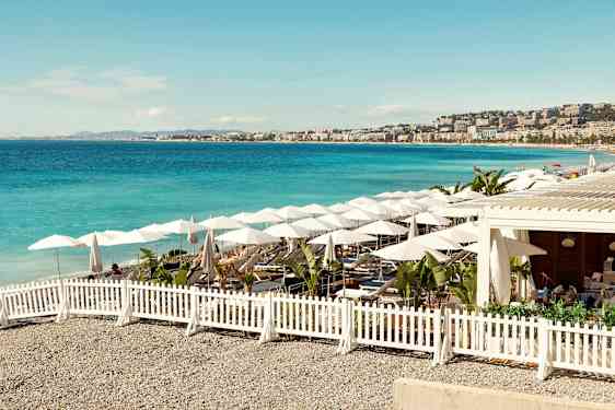 En solrik strand med blått hav, parasoller og sitteplasser omgitt av et hvitt gjerde. (AI-generert tekst)