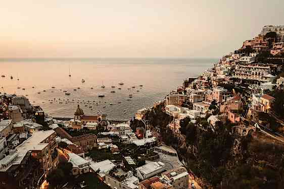 Bilde av Positano ved solnedgang med bygninger langs en skråning og havet fylt med båter. (AI-generert tekst)