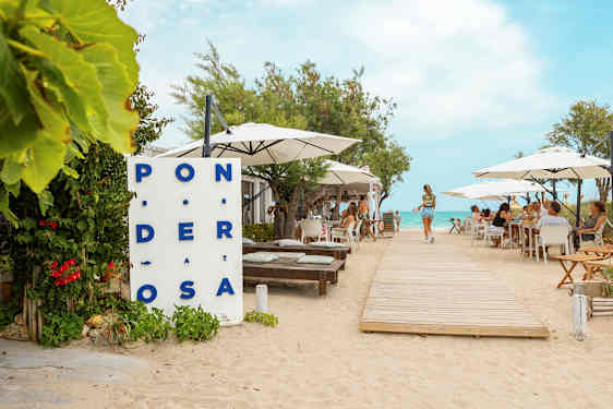 En restaurant ved stranden med bord og parasoller, en tresti fører mot vannet. (AI-generert tekst)