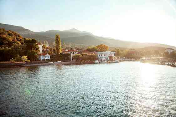 En liten landsby ligger ved en vannkant med hus og trær omgitt av fjell. Solen skinner over området. (AI-generert tekst)