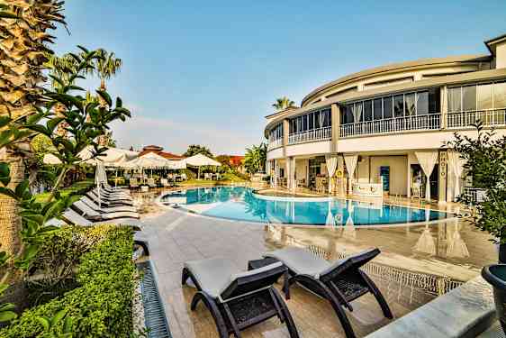 Et utendørs svømmebasseng omgitt av solsenger, parasoller og en moderne bygning. (AI-generert tekst)