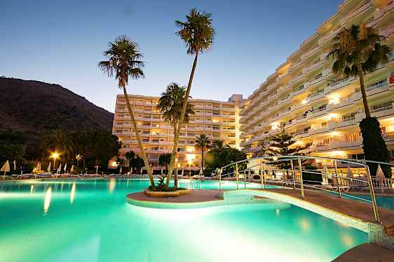 Et bilde av et hotellområde ved skumring, med et opplyst basseng, en bro og høye bygninger i bakgrunnen. (AI-generert tekst)