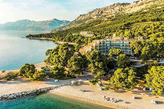 Flyfoto av en strand med parasoller, bygninger omgitt av trær og fjell i bakgrunnen. (AI-generert tekst)