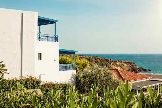 Hvit bygning med blå detaljer nær sjøen og grøntområder. (AI-generert tekst)