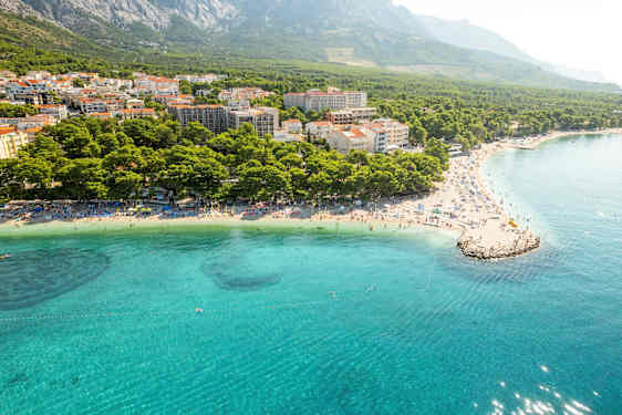 Luftbilde av en kyst med klart blått vann, strand med solsenger og tett bebyggelse omgitt av skog og fjell. (AI-generert tekst)