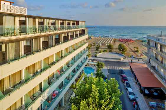 En hotellbygning med balkonger, en gate med parkering og fortau, samt en strand med solsenger og havet i bakgrunnen. (AI-generert tekst)