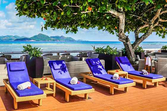 Solstoler med blå puter og sammenrullede håndklær plassert ved et hav med fjell i bakgrunnen. (AI-generert tekst)