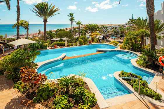 Utsikt over et basseng omgitt av grøntområder og palmer, med havet i bakgrunnen. (AI-generert tekst)