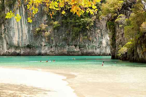 Bildet viser en strand med klart tyrkis vann omgitt av høye klippeformasjoner dekket av vegetasjon. Flere personer svømmer i vannet under en klar himmel. (AI-generert tekst)