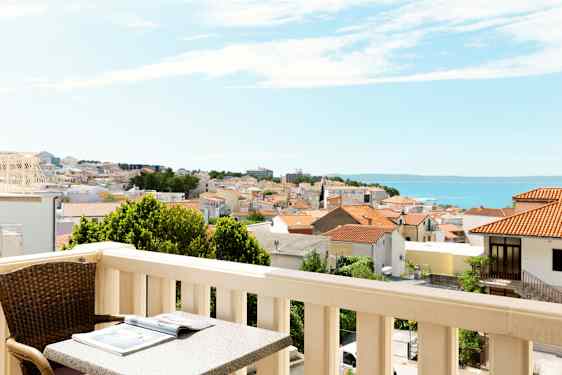Balkong med bord og stol, utsikt over en by med røde tak og havet i bakgrunnen. (AI-generert tekst)