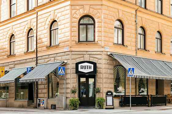 Et hjørnehus i klassisk stil med butikkens skilt Ruth over inngangen. Utstillingsvinduer med stripete markiser, gangvei og stolpeskilt er synlige i forgrunnen. (AI-generert tekst)