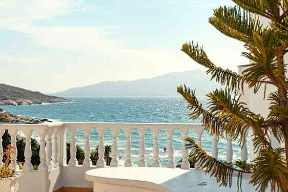 Utsikt over havet fra en terrasse med hvitt rekkverk og en del av et bord i forgrunnen sammen med en barlignende plante. (AI-generert tekst)