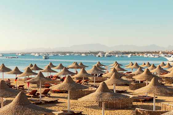 En strand med solsenger og parasoller av naturmaterialer, med sjøen og båter i bakgrunnen. (AI-generert tekst)