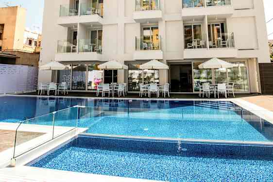 Et hotell med flere etasjer, balkonger og et basseng foran bygningen samt utendørs servering med bord og parasoller. (AI-generert tekst)