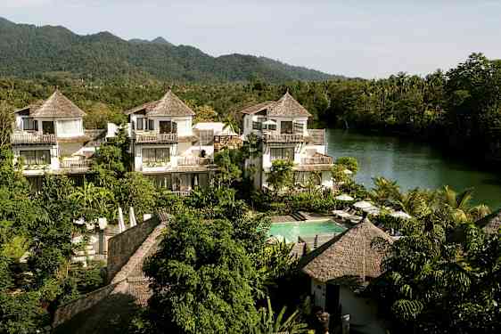 Et hotellområde med hus i tropisk stil, omgitt av grøntområder og en elv i bakgrunnen. (AI-generert tekst)