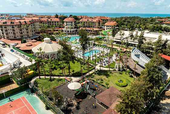 Oversiktsbilde av et feriested med hotell, bassenger, lekeplass og hager nær sjøen. (AI-generert tekst)