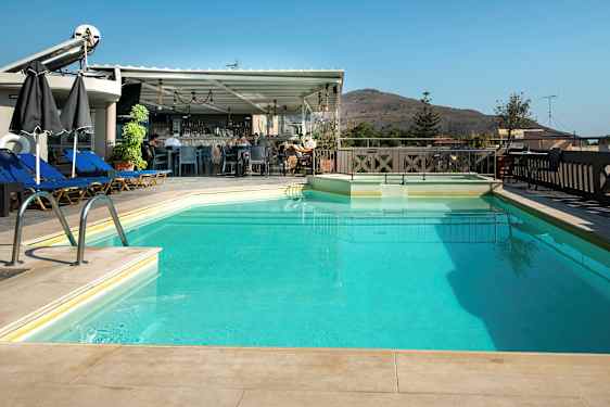 Et utendørs svømmebasseng med solsenger og parasoller ved siden av, samt en bar i bakgrunnen. Et fjell ses i det fjerne. (AI-generert tekst)