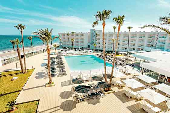 En utsikt over et feriested med et stort basseng, omgitt av solsenger og parasoller, med palmer og en bygning i bakgrunnen nær havet. (AI-generert tekst)