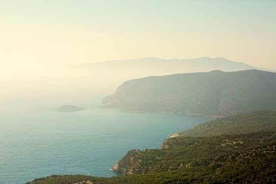 En kystlinje med bratte klipper og grønne skoger som skråner ned mot havet. Et dis dekker landskapet. (AI-generert tekst)