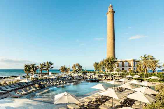 En solrik resort med basseng, solsenger og parasoller samt et fyrtårn i bakgrunnen. (AI-generert tekst)