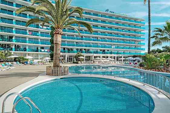 Bilde av et hotell med et basseng, omgitt av solsenger og parasoller, med palmer og grønne planter. (AI-generert tekst)